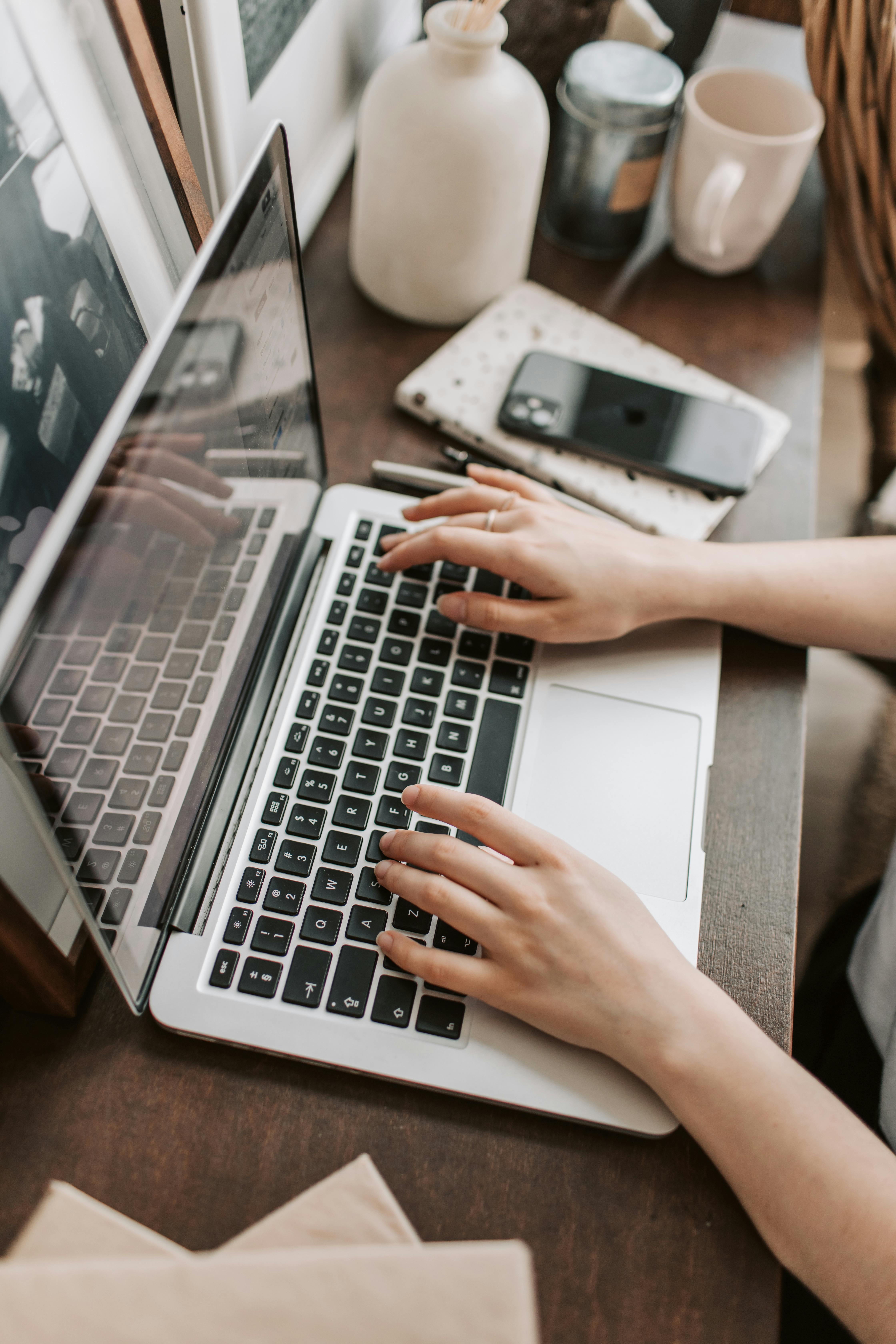 A person typing on an Apple laptop on a table. There is an iPhone to the right of the person.